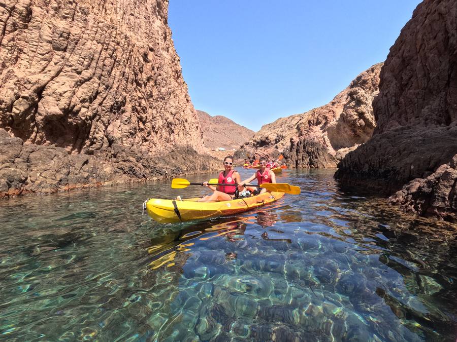 Dos personas en un kayak amarillo entre paredes de roca volcánica, sobre agua turquesa transparente, en la ruta del Corralete