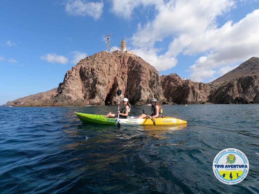 Dos personas en kayak amarillo frente al Arrecife de las Sirenas, con el faro de Cabo de Gata al fondo
